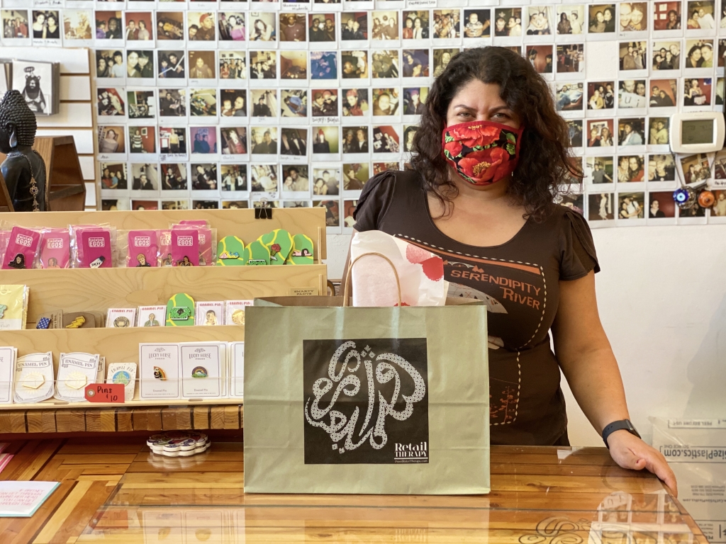 Image of Retail Therapy owner, Wazhma Samizay, wearing a facemask standing behind the counter of her store. There is a bag with a gift on the counter in front of her.